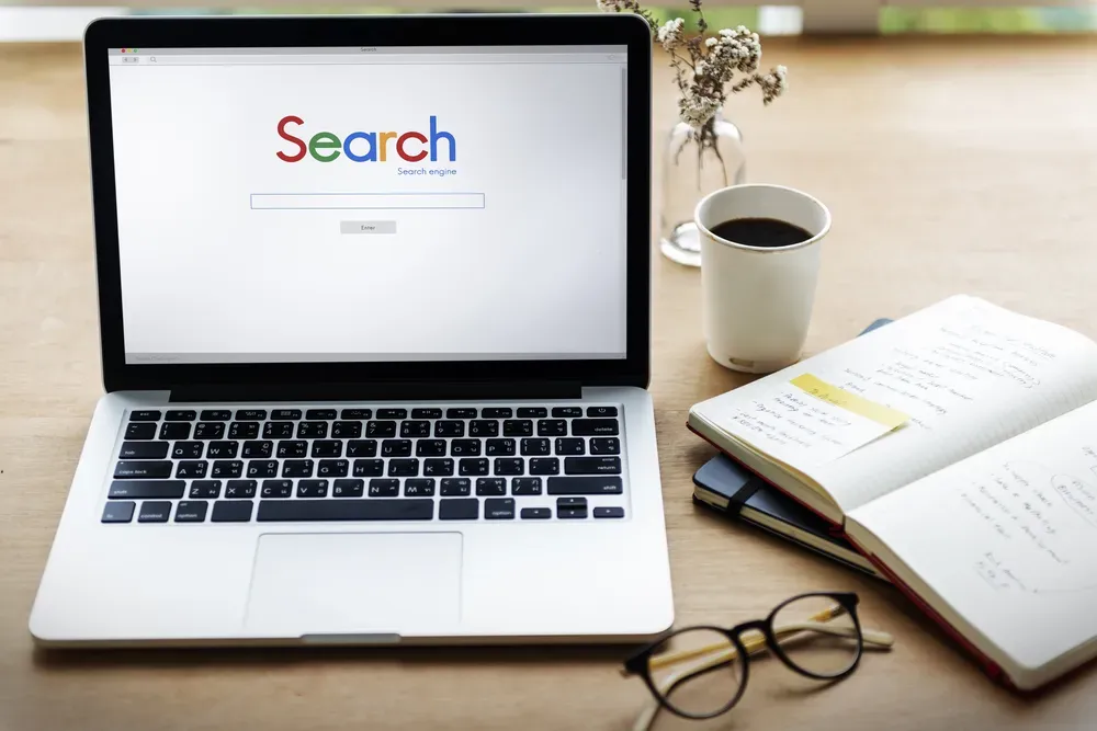A photo of a desk with a laptop with a search engine screen, a notebook, coffee and glasses.
