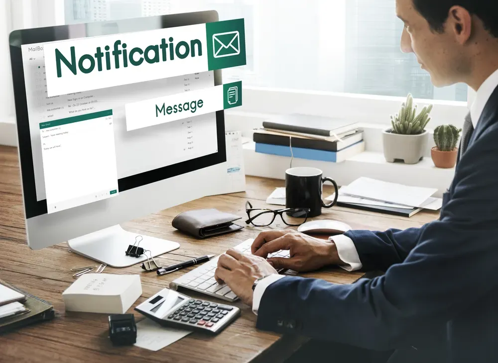 A businessman in a suit sitting at a desk and computer with large notification infographics on the monitor screen.