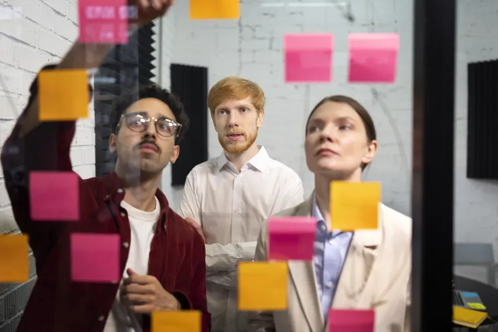 Picture of 3 people thinking on colorful sticky notes stuck on a transparent wall.