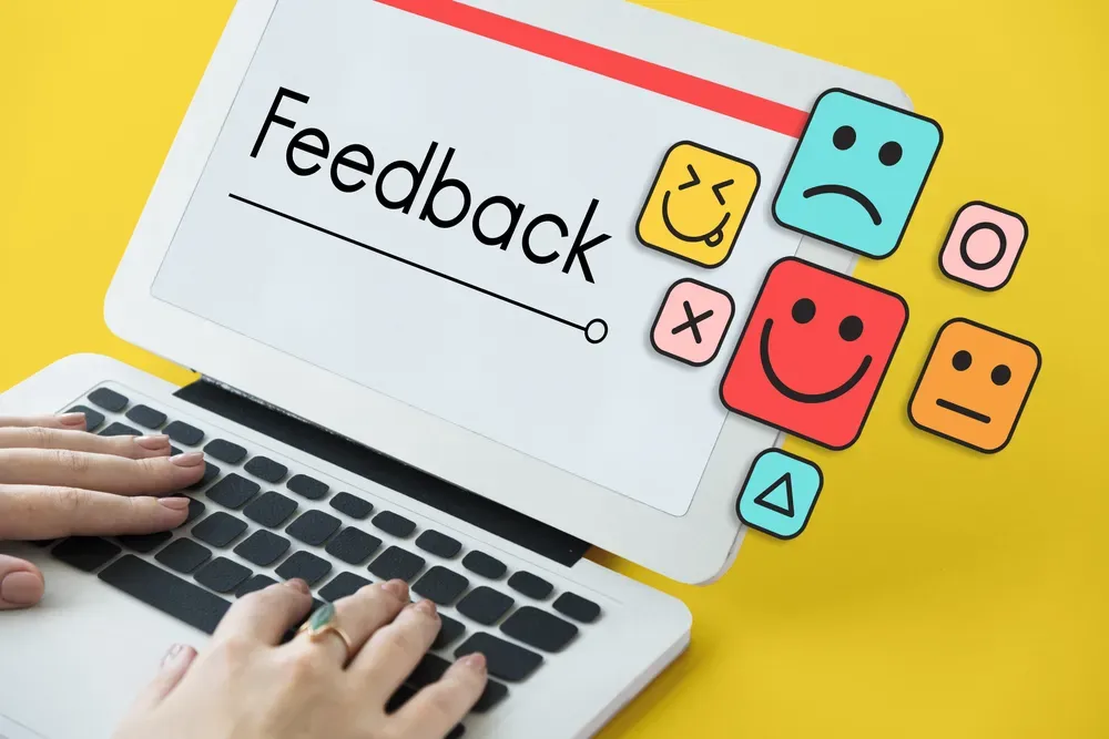 A toy laptop with female hands on it and the word feedback on the screen and infographics of emoticons next to the screen.