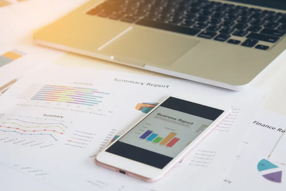Photo of a desk with sheets of paper with data, a phone showing infographics with data and a laptop.