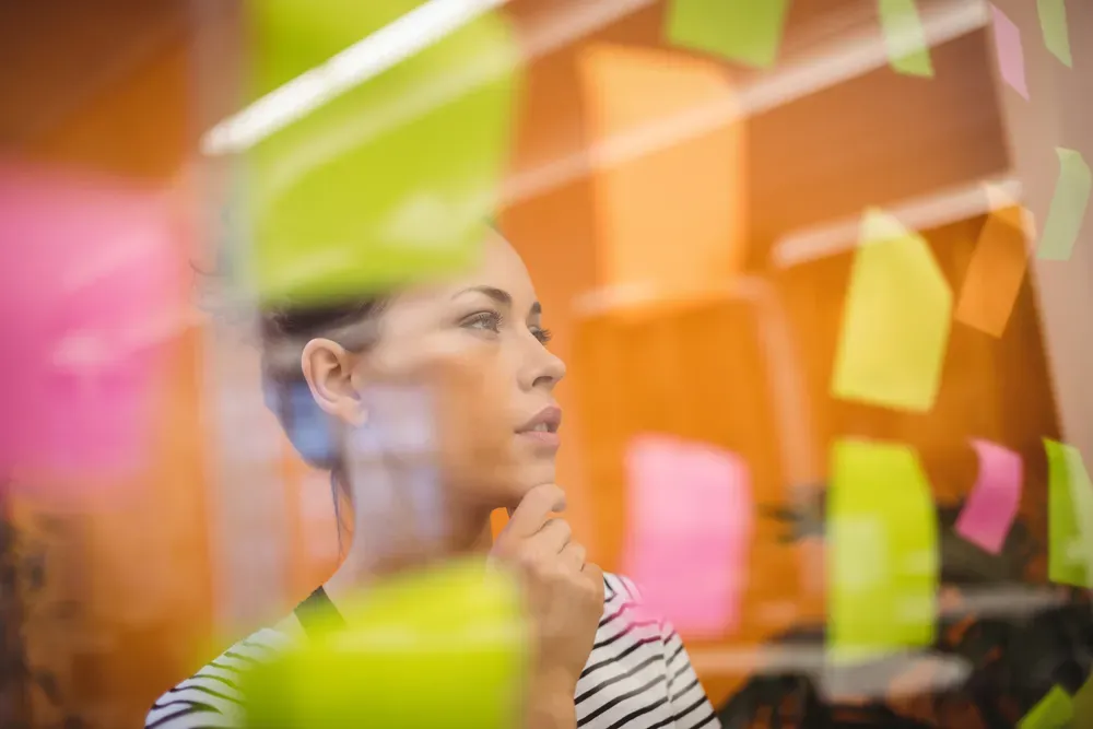 A woman standing and thinking over a transparent window with small colorful notes glued to it.